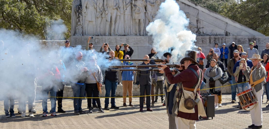 Eine Gruppe Männer in historischer Kleidung schießt bei einer Vorführung mit Langgewehren auf einem Platz, dahinter Zuschauende vor einem Denkmal.