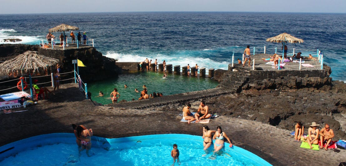 Personen baden in Meerwasserbecken an einer Felsküste mit dunklem Vulkangestein.