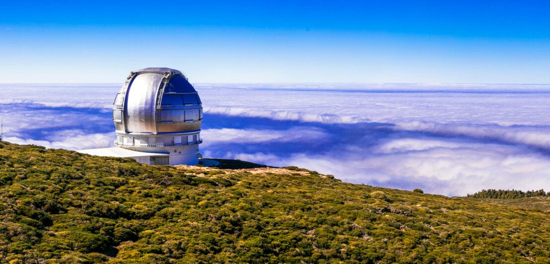 Ein kugelförmiges Observatorium auf einem grünen Bergplateau über der Wolkendecke.