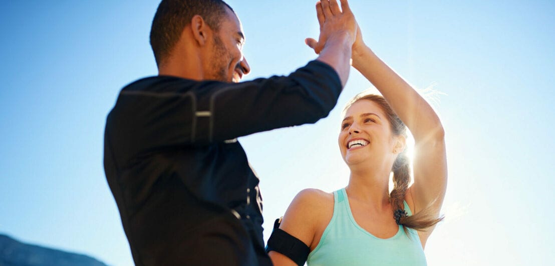 Ein Mann und eine Frau in Sportkleidung schlagen gut gelaunt mit den Händen ein, im Hintergrund blauer Himmel.