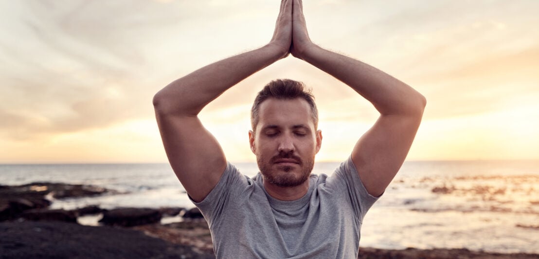 Ein Mann führt mit geschlossenen Augen eine Yogaübung mit über dem Kopf gefalteten Händen am Strand aus.
