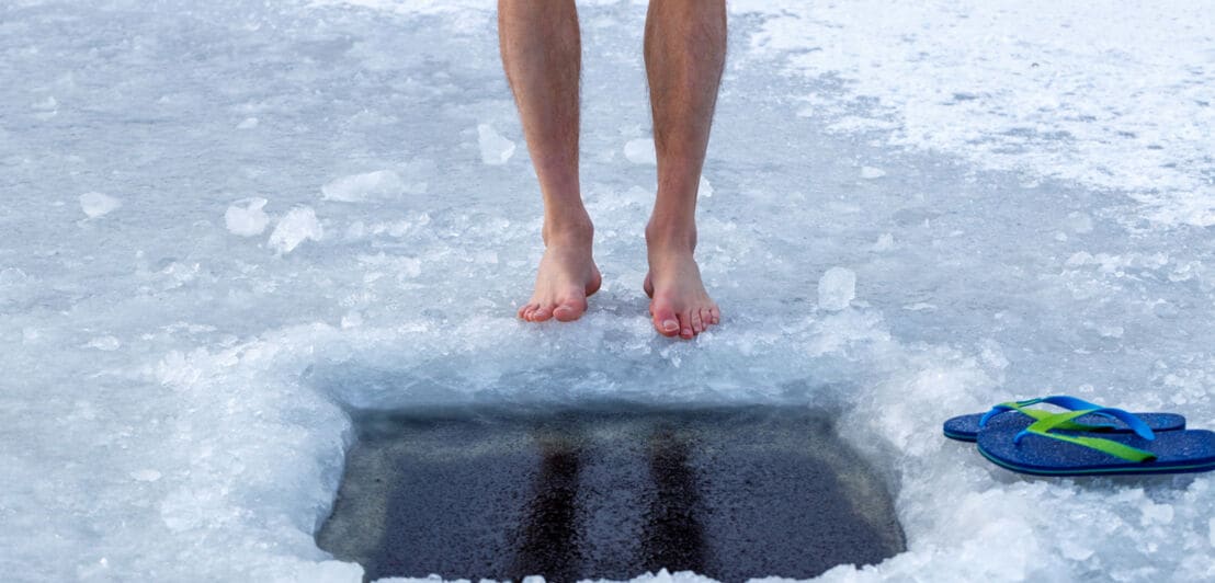 Zwei nackte Beine auf einem zugefrorenen See vor einem aufgebohrten Wasserloch, daneben zwei Badeschlappen.