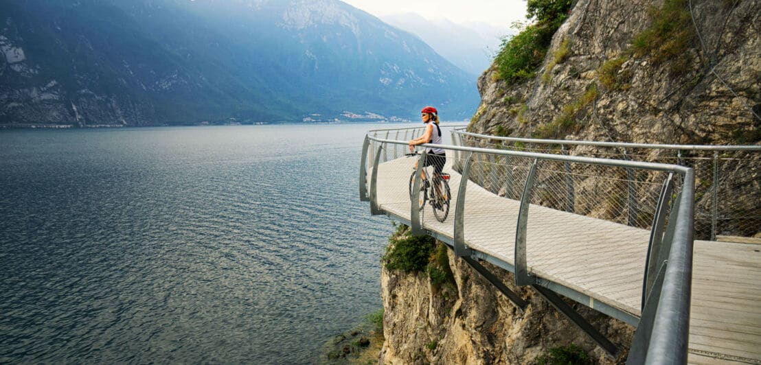 Eine Radfahrerin steht am Geländer eines Radweges an einem Felshang oberhalb eines Sees.