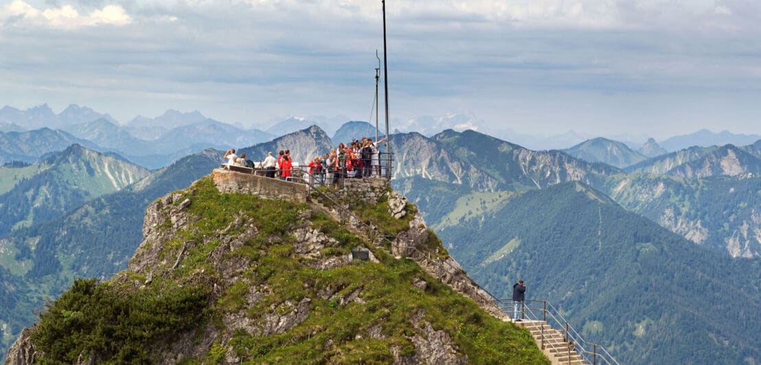 Personen auf einer Aussichtsplattform auf einem Berggipfel vor Alpenpanorama.