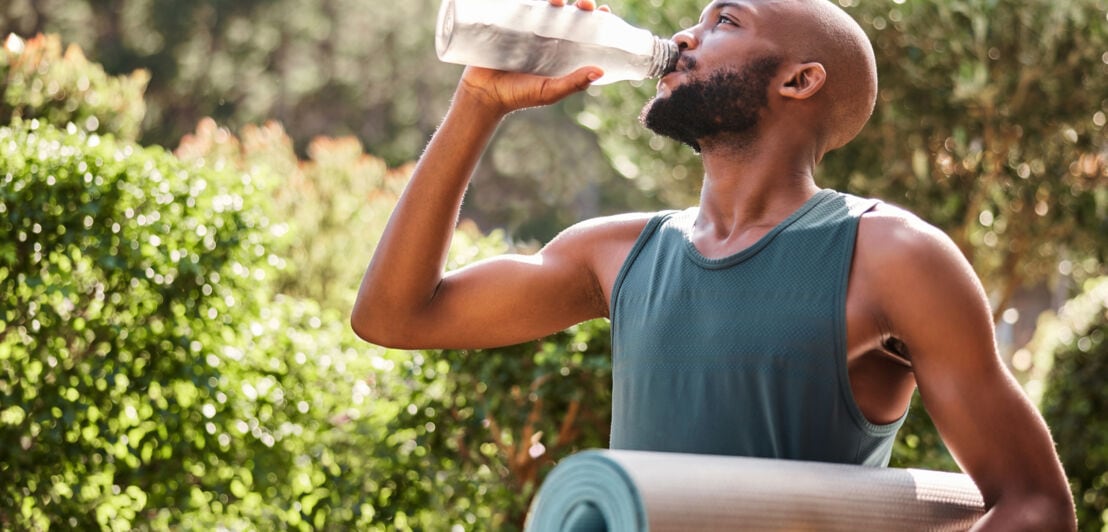 Ein junger, durchtrainierter Mann mit Yogamatte unter dem Arm trinkt aus einer Wasserflasche draußen in der Natur.