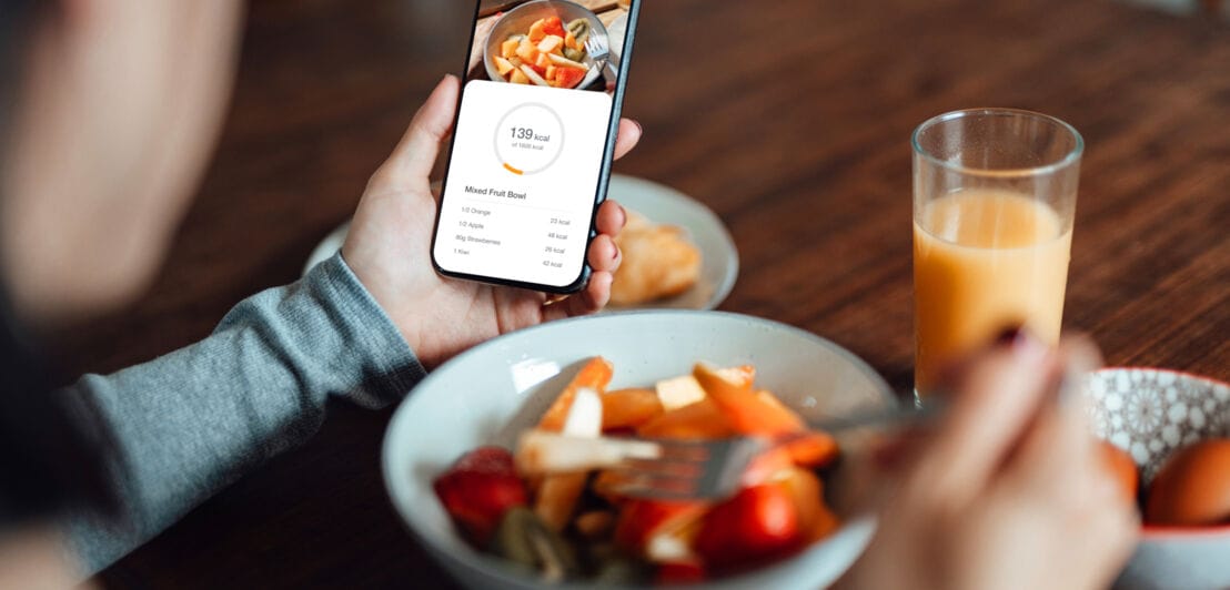 Eine Person schaut beim Essen eines Gemüsegerichtesauf das Display ihres Smartphones mit Kalorienzähler.