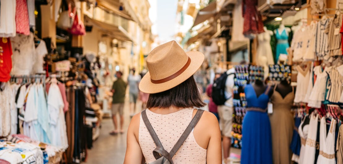 Rückansicht einer Frau mit Strohhut, die an einem Urlaubsort durch eine Einkaufsstraße läuft.
