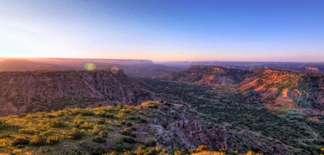 Landschaftspanorama eines weitläufigen Canyons mit roten Felsen und grüner Vegetation bei Sonnenuntergang.