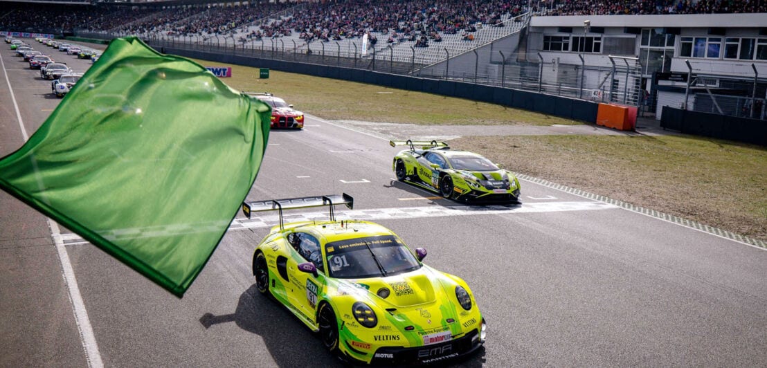 Ein Autorennen mit Zuschauern auf Tribünen, im Vordergrund eine grüne Flagge vor einem gelben Porsche.