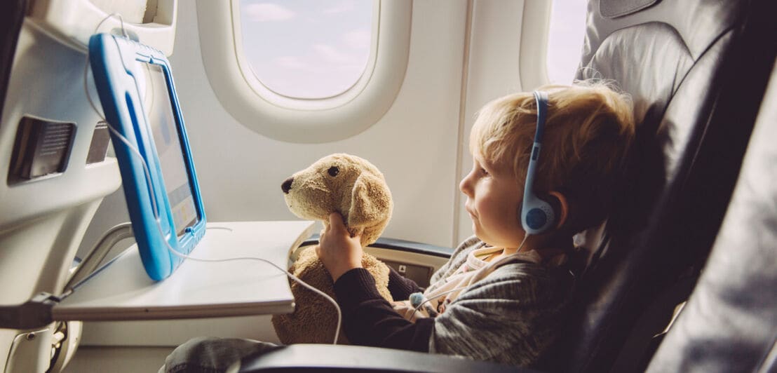 Ein kleiner Junge mit Kopfhörern in einem Flugzeugsitz schaut mit seinem Plüschhund einen Film auf einem Tablet.