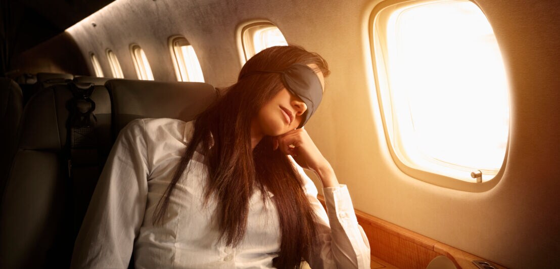 Eine Frau mit langen Haaren schläft mit Schlafmaske in der Business Class im Flugzeug.