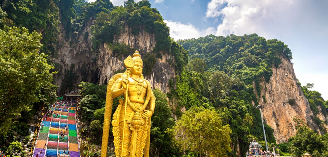 Tempelanlage mit goldener Buddhastatue vor einer Felswand mit Höhle, zu der eine bunte Treppe hinaufführt.