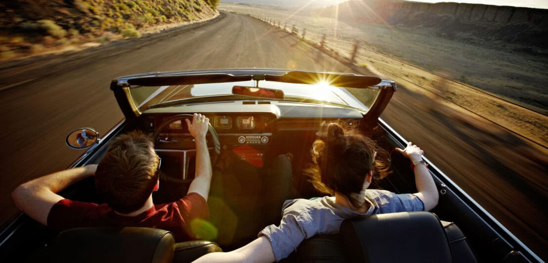 Rückansicht eines Paares in einem offenen Cabriolet während der Fahrt auf einer Straße durch eine Wüstenlandschaft bei Sonnenuntergang. 