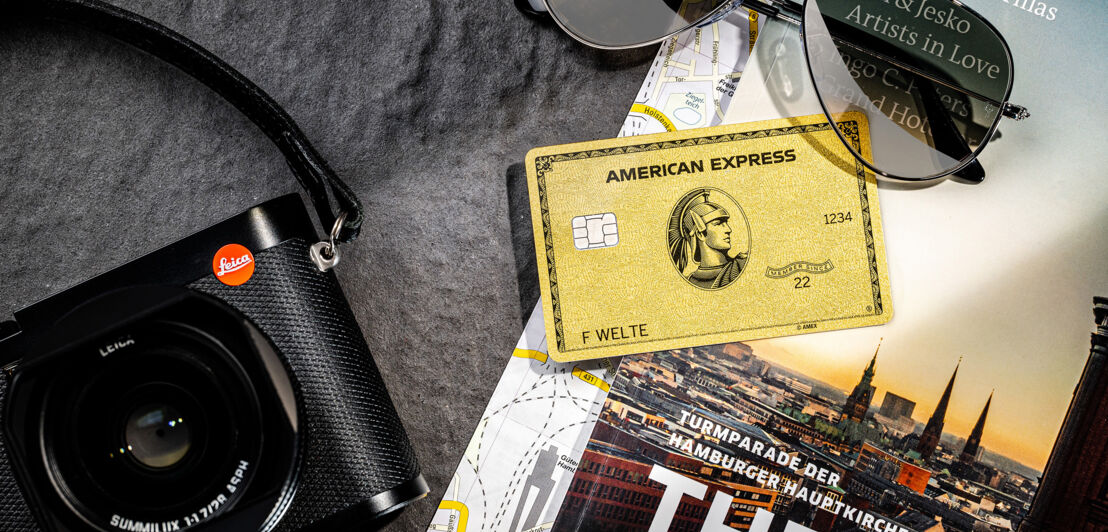 Eine goldene Kreditkarte liegt neben einer Kamera auf einem Reiseführer.