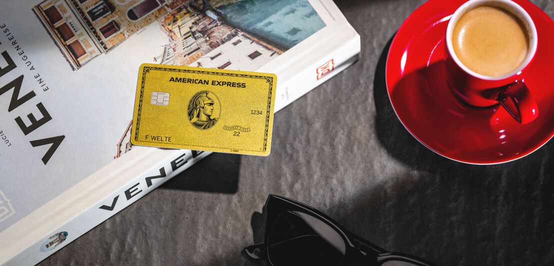 Eine American Express Gold Card liegt auf einem Buch neben einem Espresso.