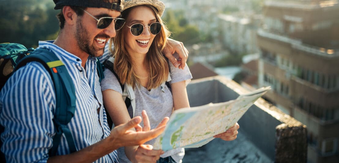 Ein junges Paar mit Rucksäcken und Sonnenbrillen blickt auf einen Stadtplan in einer Stadt.