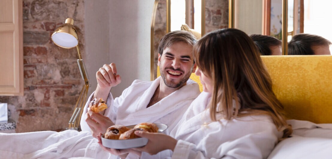 Ein gut gelauntes Paar in weißen Bademänteln mit Croissants in den Händen liegt auf einem Bett.