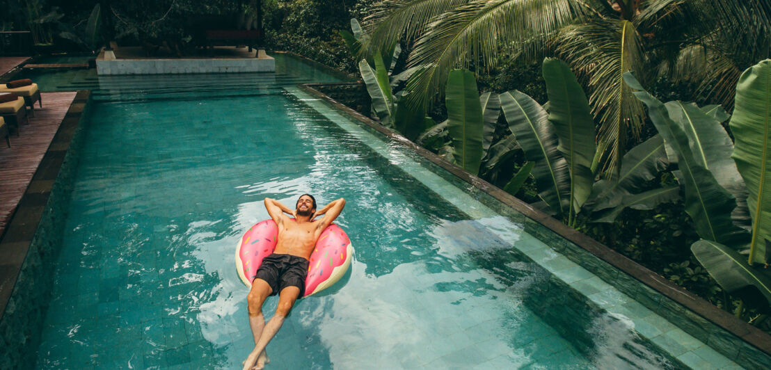 Ein Mann schwimmt entspannt rücklings auf einer pinken Luftmatratze in einem Hotelpool in tropischer Umgebung.