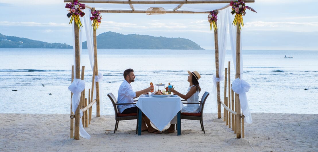 Ein Paar sitzt beim Essen an einem Tisch unter einem dekorierten Baldachin am Strand.