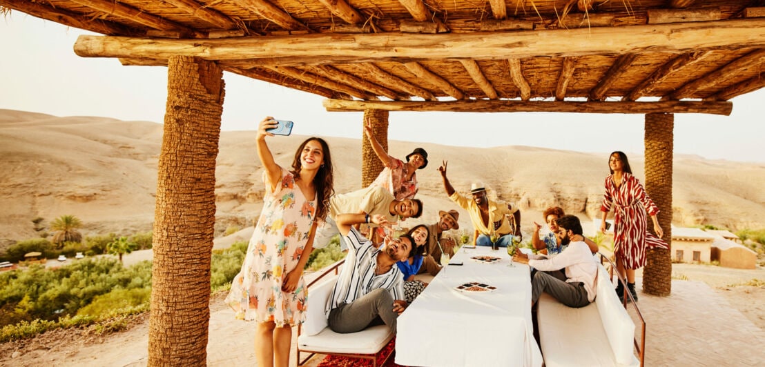 Eine Frau macht ein Selfie vor einer Personengruppe an einem Tisch in einem Wüstencamp.
