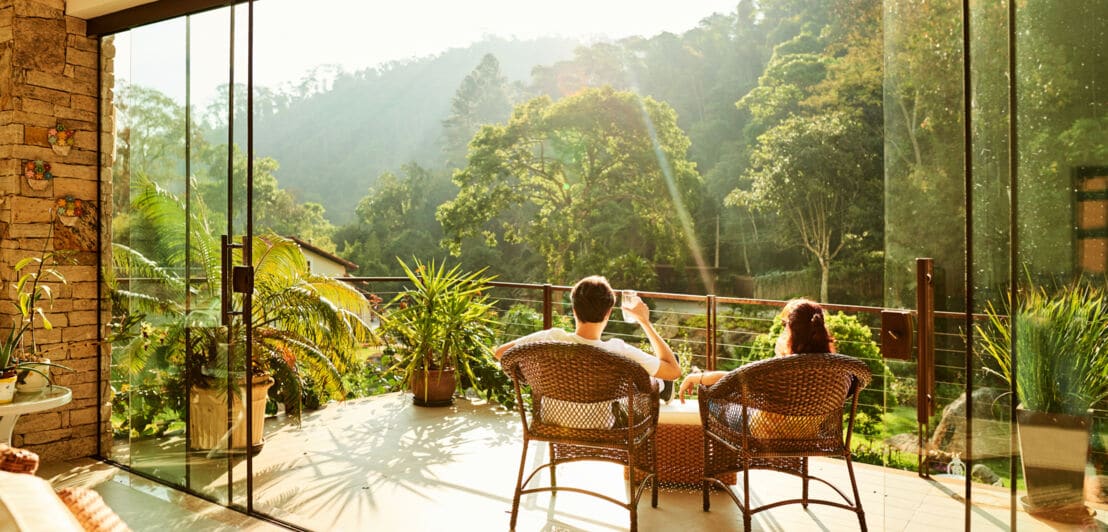 Rückansicht eines Paares in Korbstühlen, das mit Getränken auf einer Balkonterrasse sitzt und in einen Dschungel blickt bei Sonnenschein.