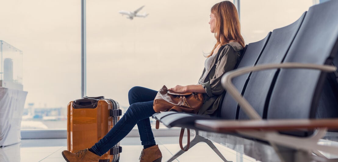 Eine Frau mit Koffer sitzt alleine im Wartebereich eines Flughafengates und blickt hinaus aufs Rollfeld