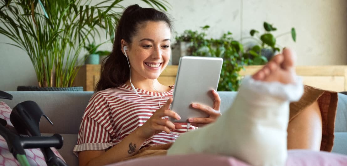 Eine junge Frau auf einem Sofa schaut lächelnd auf ihr Tablet, während sie ihr eingegipstes Bein hochgelegt hat