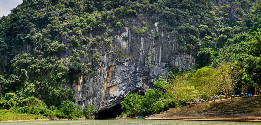 Eine Landschaft mit einem Fluss im Vordergrund und einer Felswand im Hintergrund, in die ein Höhleneingang führt.