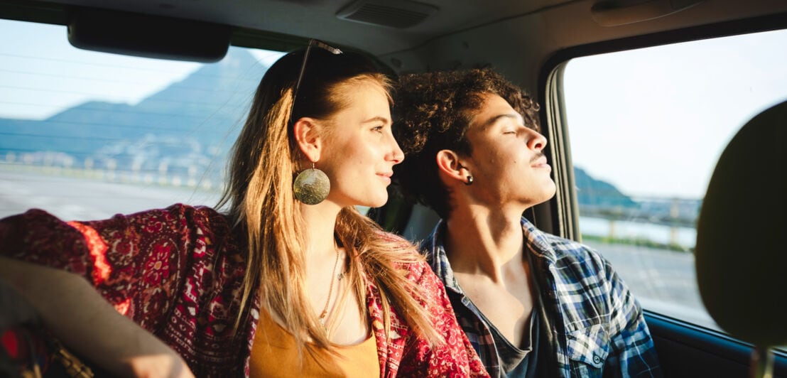 Ein junges Paar auf dem Rücksitz eines Autos blickt aus dem Fenster bei warmem Sonnenlicht.