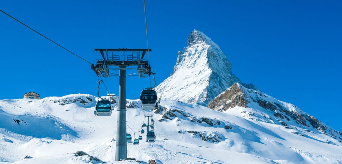 Berggondel mit schneebedeckter Piste, Matterhorn und blauem Himmel im Hintergrund.