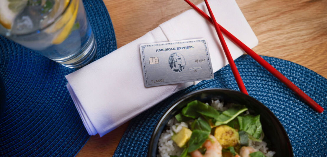 Eine American Express Platinum Card liegt auf einer weißen Serviette, daneben liegen rote Stäbchen sowie ein Teller mit asiatischem Essen.