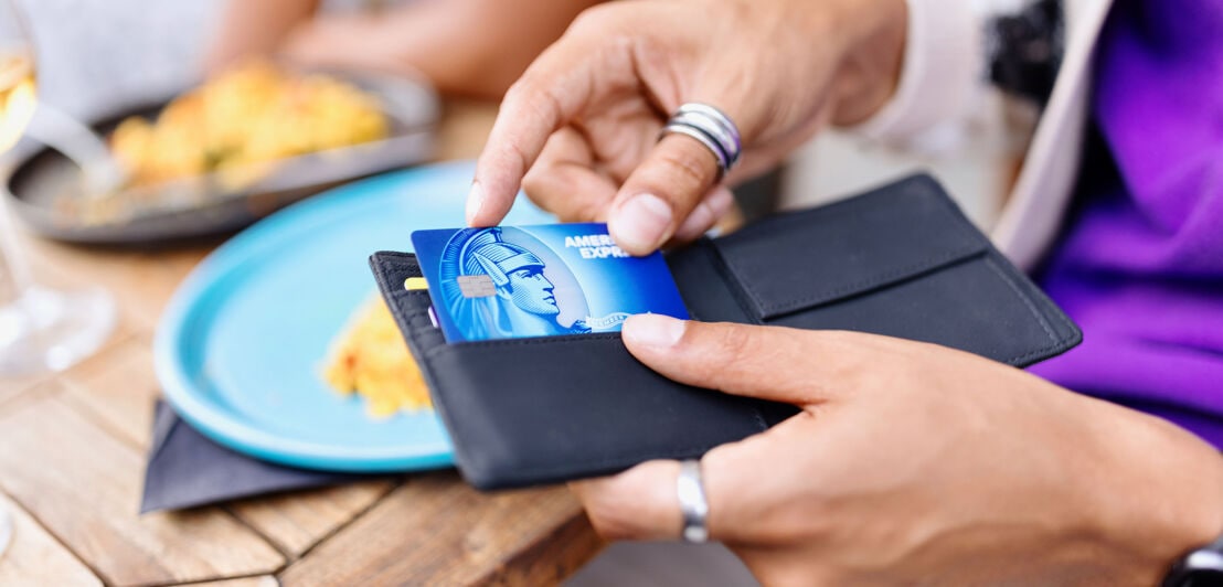 Eine Person greift nach einer Blue Card von American Express in einem Portemonnaie an einem Esstisch.