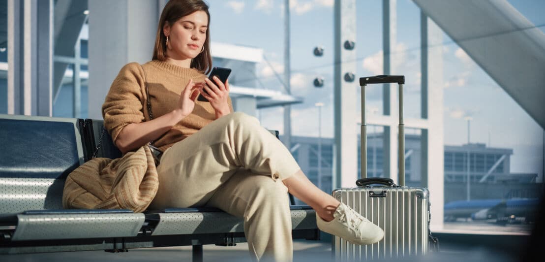 Eine Frau sitzt mit überschlagenen Beinen auf einer Sitzreihe in einer Wartehalle am Flughafen und hält ein Smartphone in der Hand. Neben ihr steht ein silberner Koffer mit ausgefahrenem Griff.