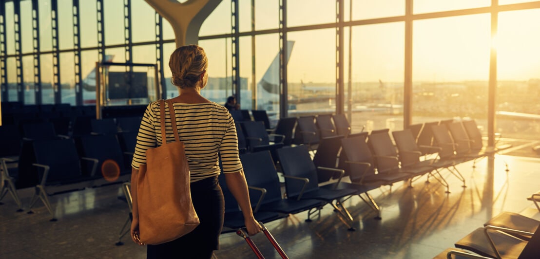 Rückenansicht einer Frau, die mit einen Koffer durch eine leere Wartehalle am Flughafen geht, im Hintergrund geht die Sonne unter.
