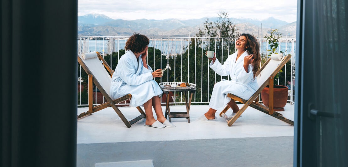 Zwei Freundinnen in weißen Bademänteln unterhalten sich bei Kaffeetrinken auf einem Balkon mit Blick auf einen See.