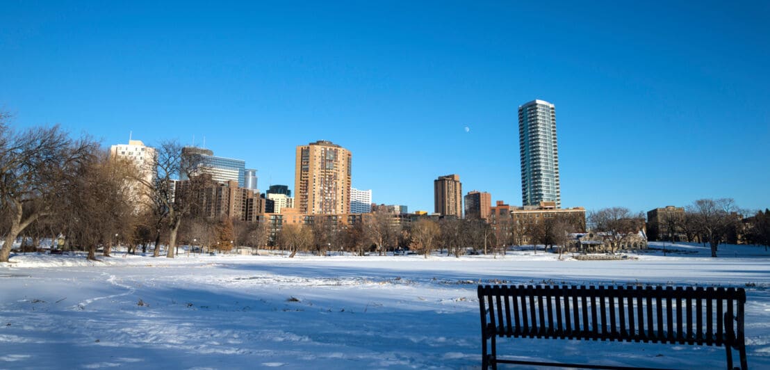 Eine Sitzbank in einem Park in Minneapolis im Winter. Im Hintergrund die Skyline der Stadt bei Sonnenschein.