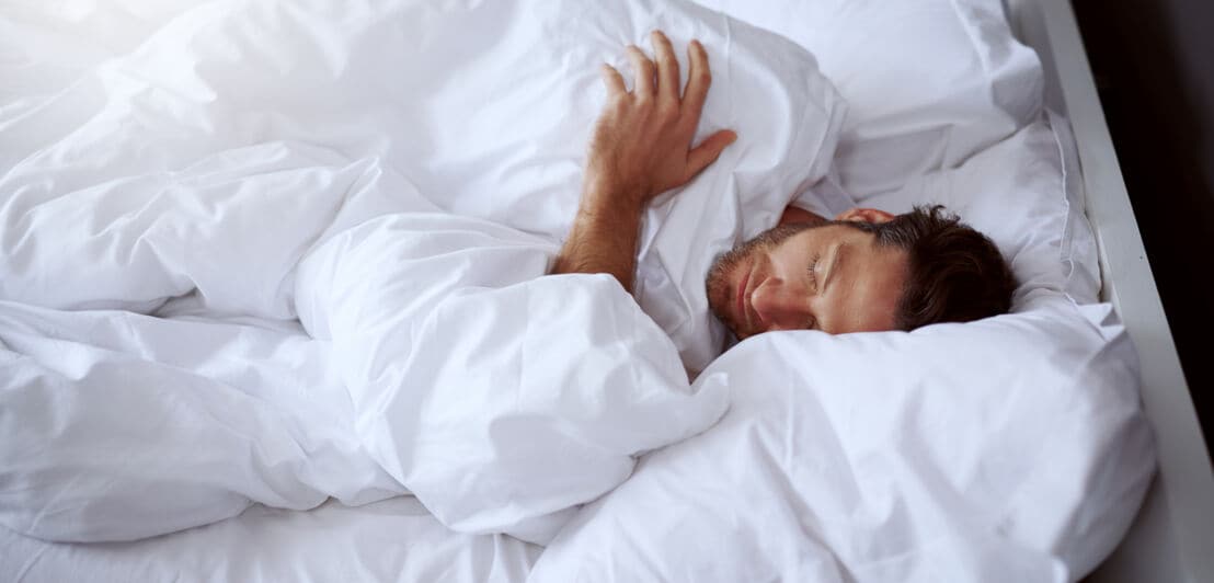 Ein Mann, der in einem Bett mit weißer Bettwäsche und Laken schläft.