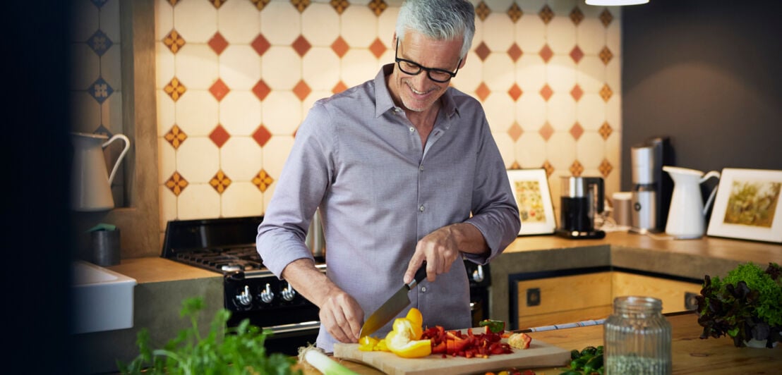 Ein lächelnder älterer Mann, der in einer Küche auf einem Küchenbrett eine Paprika schneidet.