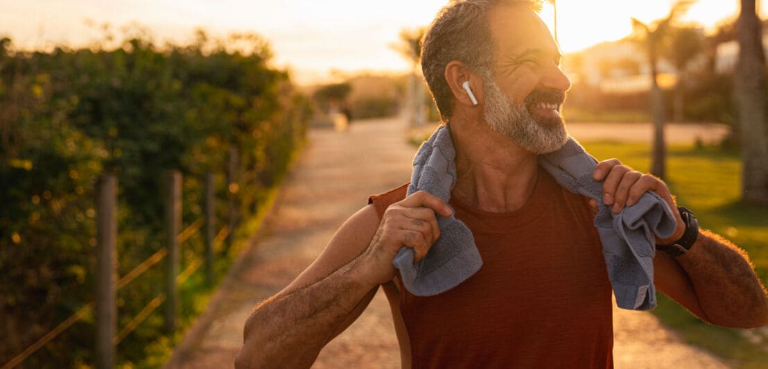Ein lächelnder älterer Mann, der sich im Freien bei Sonnenuntergang aufhält und kabellose Kopfhörer trägt. Er trägt ein Handtuch um den Hals hält dieses mit beiden Händen fest.
