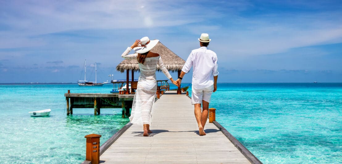 Paar in weißer Sommerkleidung geht auf den Malediven über einen Steg auf einen Anleger zu, Rückansicht