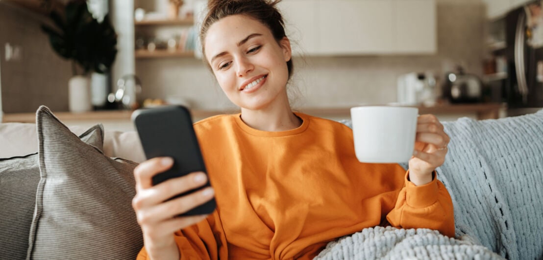 Eine junge Frau sitzt lächelnd mit Smartphone und Kaffeetasse auf einem Sofa.