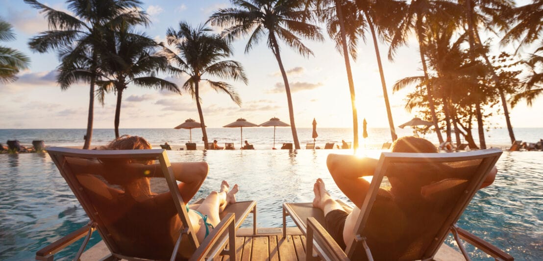 Rückansicht von zwei Personen auf Sonnenliegen vor einem palmengesäumten Hotelpool am Meer.