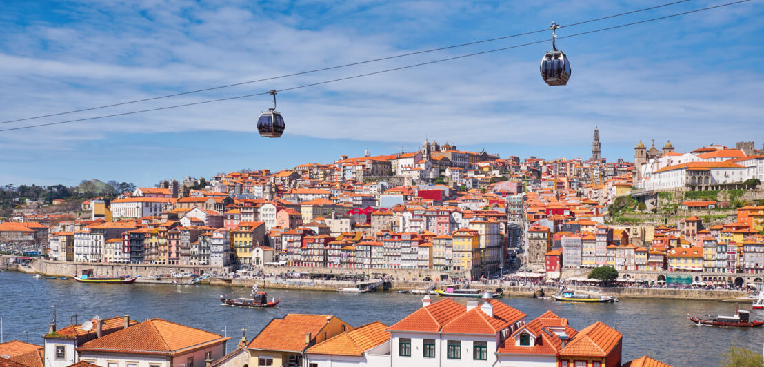 Eine Seilbahn führt über das Douro-Ufer in Porto. Im Hintergrund liegen auf einem Hügel viele bunte Häuser der Altstadt.