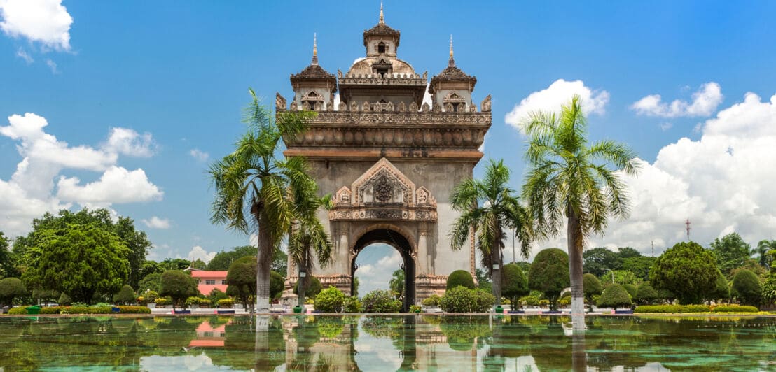 Das Patuxai-Denkmal in Vientiane, Laos. Davor Wasser mit Spiegelungen, blauer Himmel mit Wolken.