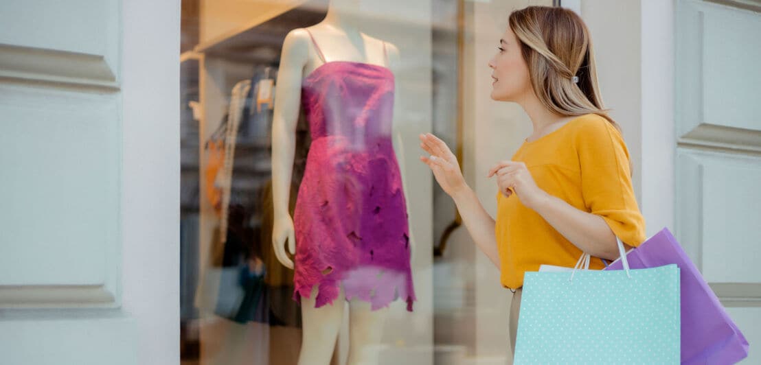 Eine Frau mit Einkaufstüten betrachtet ein pinkes Kleid im Schaufenster einer Modeboutique.