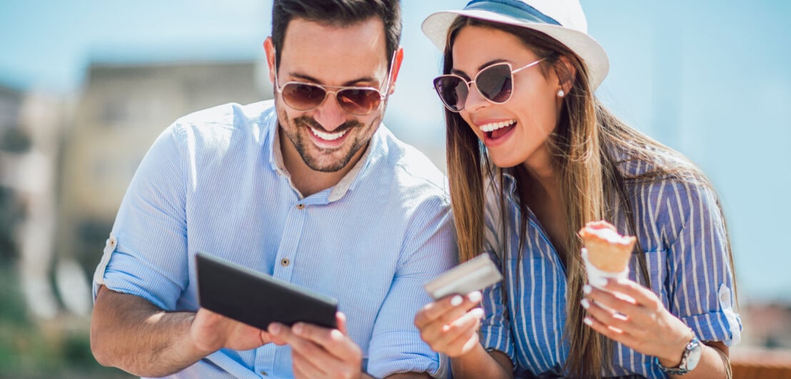 Ein fröhliches Paar in Sommerkleidung mit Sonnenbrillen schaut auf ein Tablet, sie hält eine Eiswaffel und eine silberne Kreditkarte in den Händen.