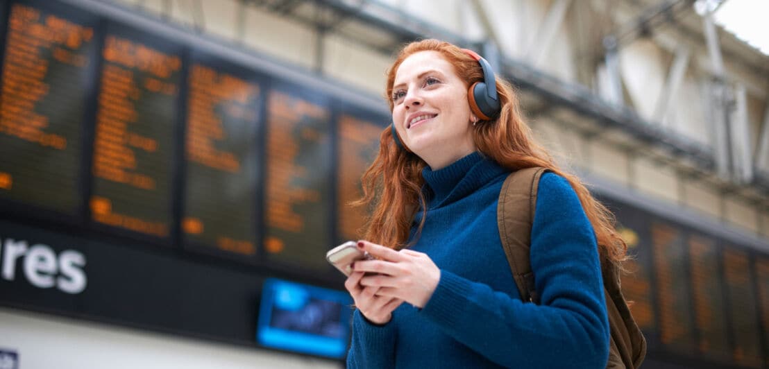 Eine junge Frau mit Kopfhörern und Smartphone steht lächelnd vor einer Abflugtafel in einem Flughafenterminal.