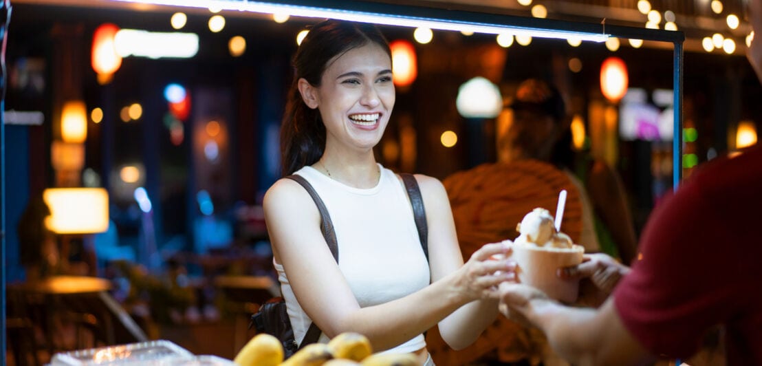 Eine junge Frau kauft Streetfood an einem erleuchteten Marktstand bei Nacht.