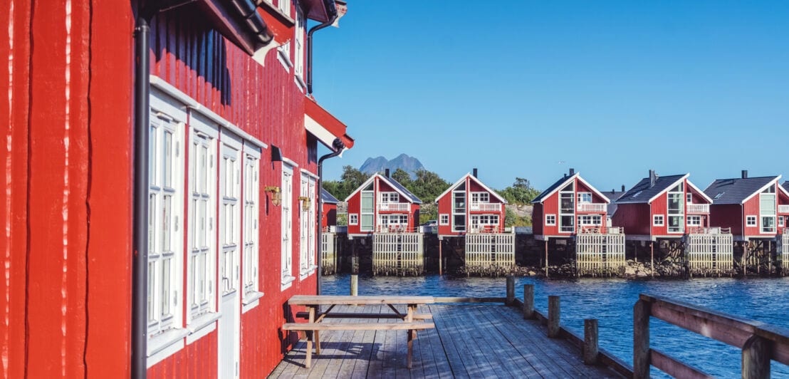 Eine Reihe roter Fischerhütten auf den Lofoten in Norwegen. Im Vordergrund ist eine Hütte mit Terrasse, Sitzbank und Tisch im Fokus.