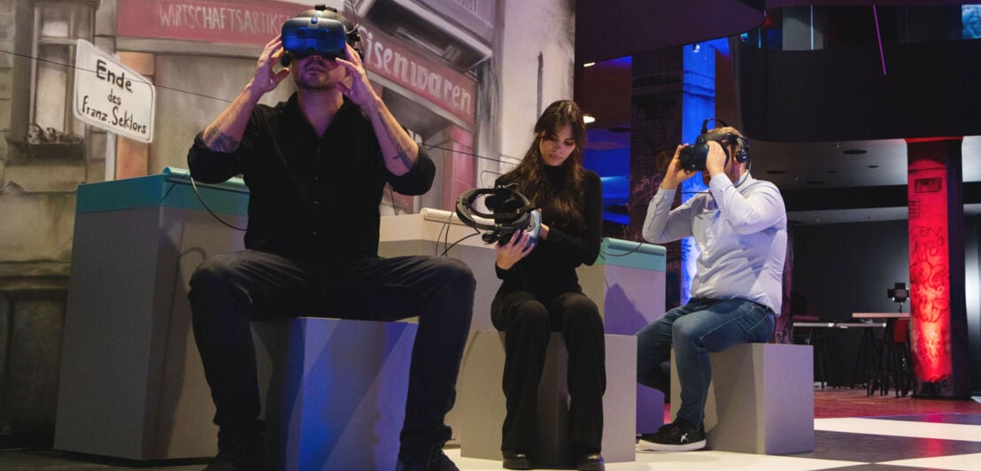 Drei Personen mit VR-Brillen sitzen auf Hockern.